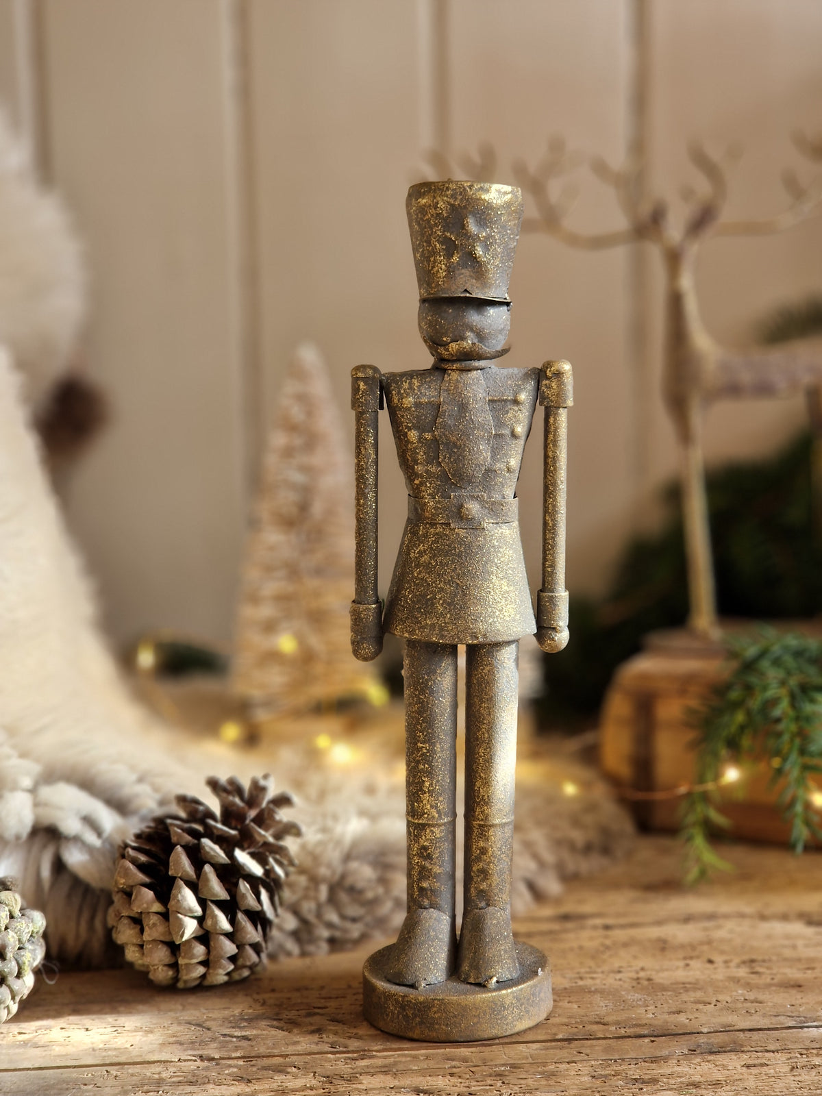 A distressed metallic gold and bronze nutcracker figurine standing on a rustic wooden surface, surrounded by holiday decorations including pinecones, a small Christmas tree, and warm string lights.