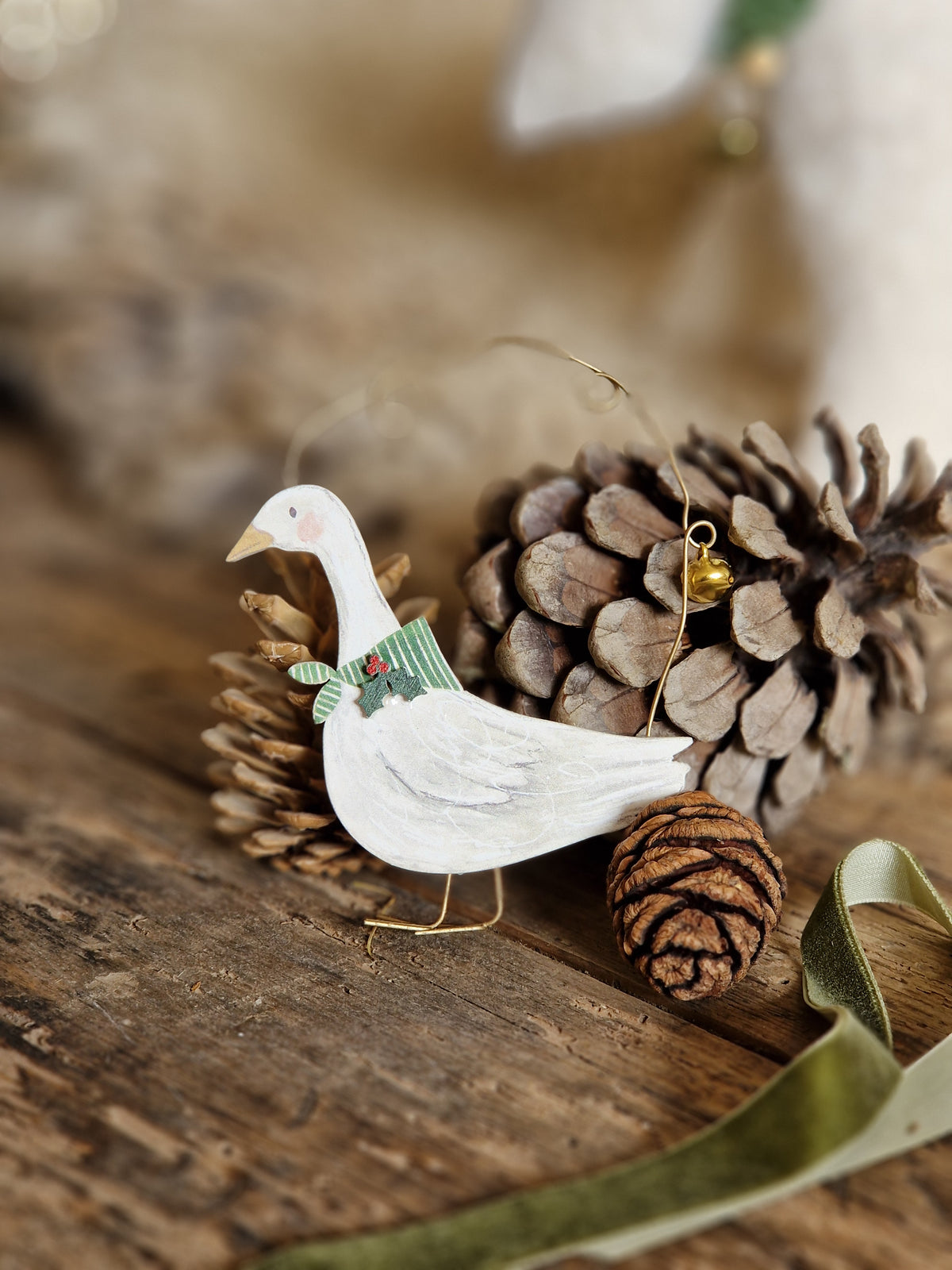 A white painted goose ornament with a green scarf and small red accent, hanging near pinecones and a wooden surface.