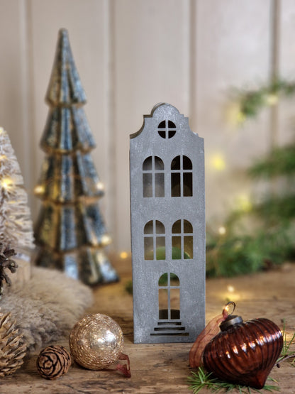A small gray metal house-shaped ornament with arched windows stands on a wooden surface, styled with holiday decorations including pinecones, a gold ornament, a dark amber ornament, and a blurred metallic tree ornament in the background.
