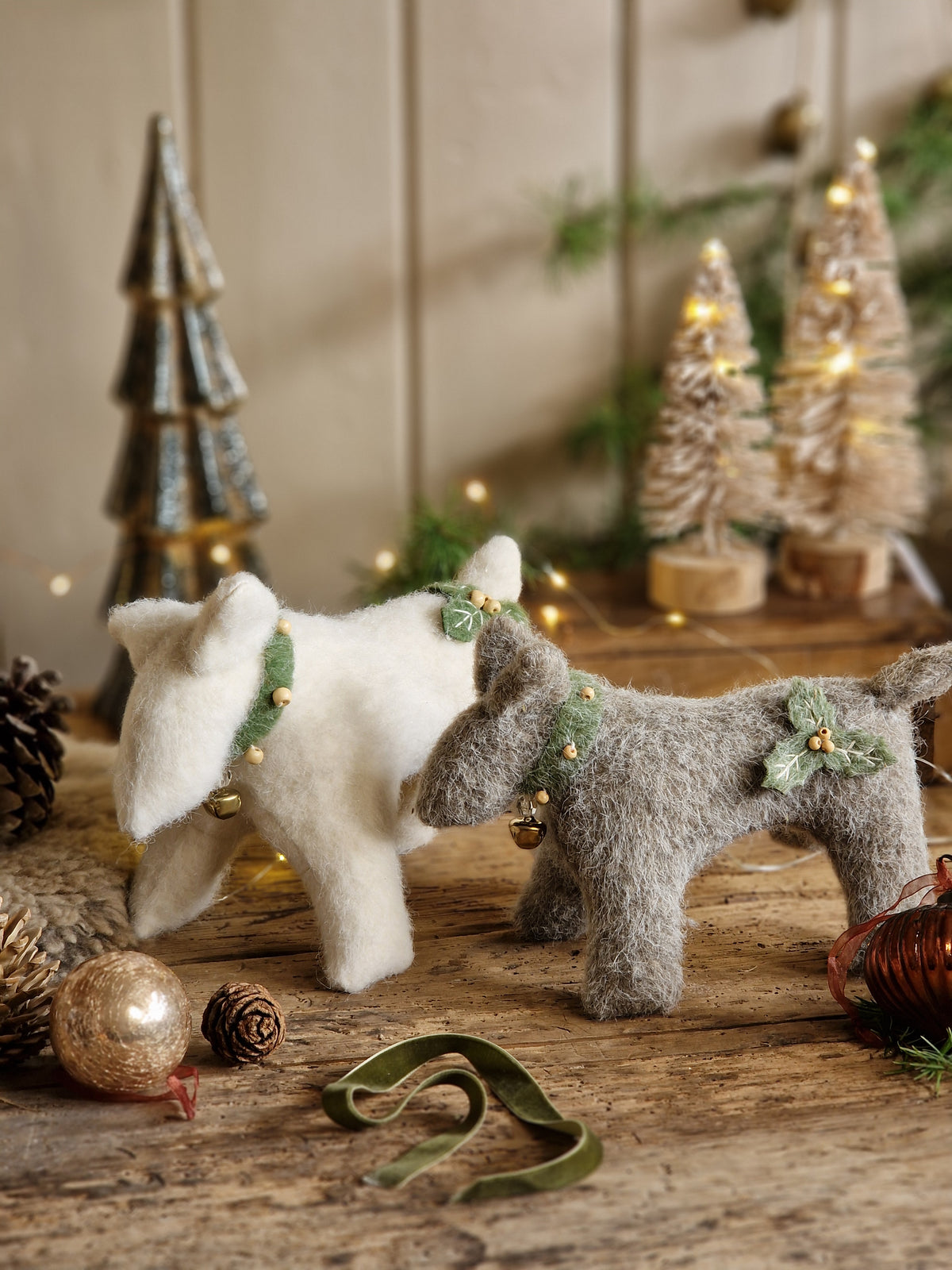 Two small felted wool dog ornaments, one white and one gray, decorated with green holly and pine branches and small gold bells, displayed on a rustic wooden surface with Christmas decorations in the background.