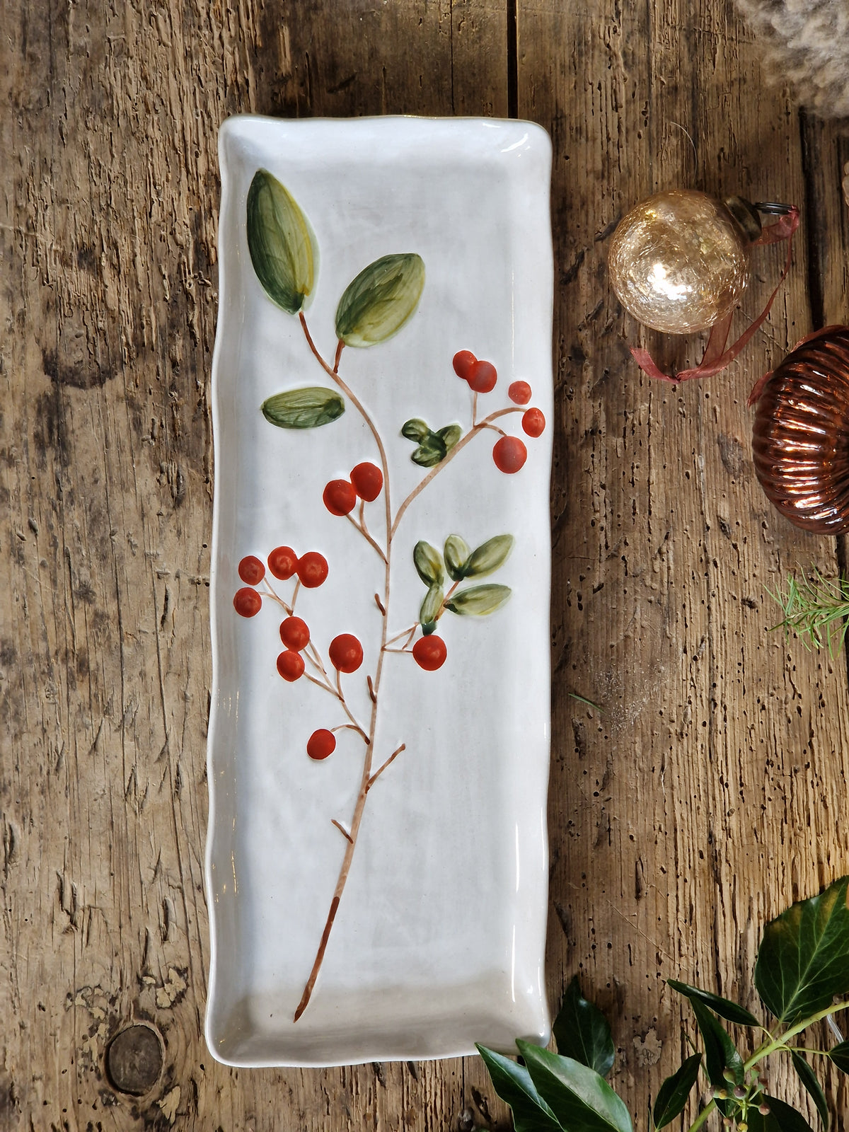A rectangular white ceramic serving platter decorated with a painted holly branch featuring green leaves and red berries, displayed on a rustic wooden tabletop with holiday ornaments.